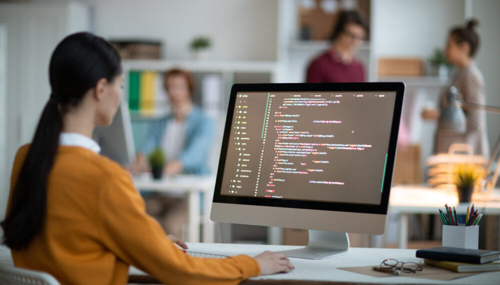 Young female programmer in casualwear sitting by desk in front of computer screen and analyzing coded data